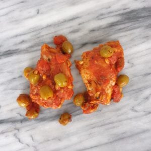 Twee stukken kip in tomatensaus met olijven en pijnboompitten op een marmeren ondergrond